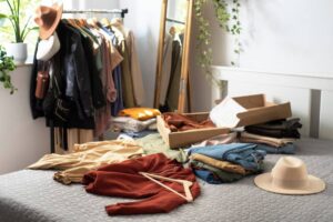 A cozy room with a bed displaying folded clothes, a box of new garments, a hat, and a clothing rack in the background.
