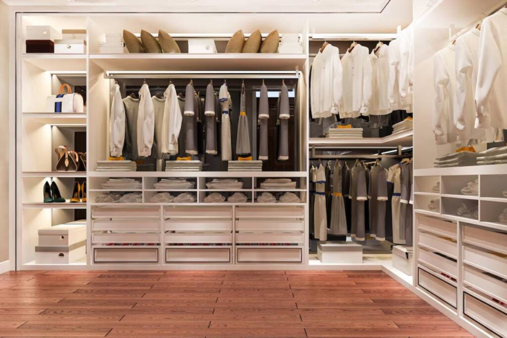Stylish organized closet with neatly hung clothes, stacked towels, and shoes on display, featuring warm lighting and wooden flooring.