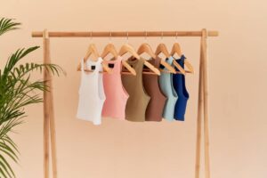 A wooden rack holds six colorful tank tops in white, pink, brown, green, blue, and navy, against a soft peach background.