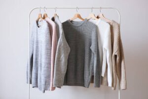 A rack displaying five cozy knit sweaters in grey, pink, white, and beige, set against a light neutral background.