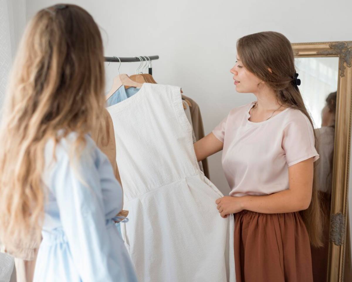 Two women are in a fashion boutique, one holding a white dress while the other looks on, surrounded by clothing on hangers.