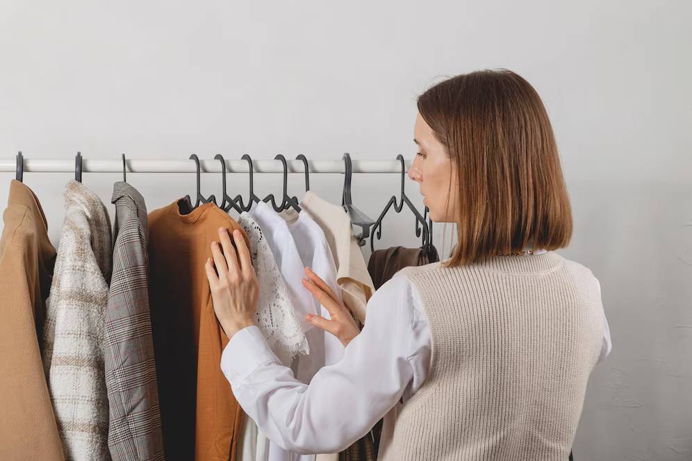 A person examines a selection of stylish clothing on hangers, including sweaters, blouses, and dresses in neutral tones.