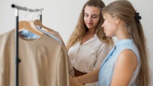 Two women are browsing clothes on hangers in a bright, minimalistic setting, engaging in conversation about their choices.