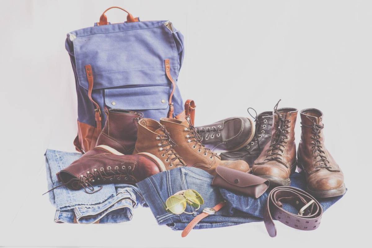 A blue backpack surrounded by various boots, jeans, sunglasses, and a belt, showcasing a laid-back, travel-ready outfit.
