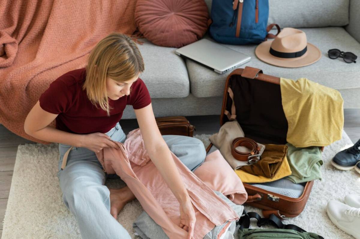 A person is packing a suitcase in a cozy living room, surrounded by clothing, a laptop, a backpack, and other travel essentials.