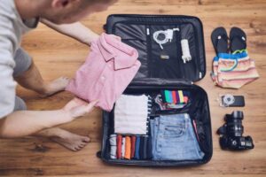 A person folding a pink polo shirt while packing a suitcase with clothes, a camera, and accessories on a wooden floor.