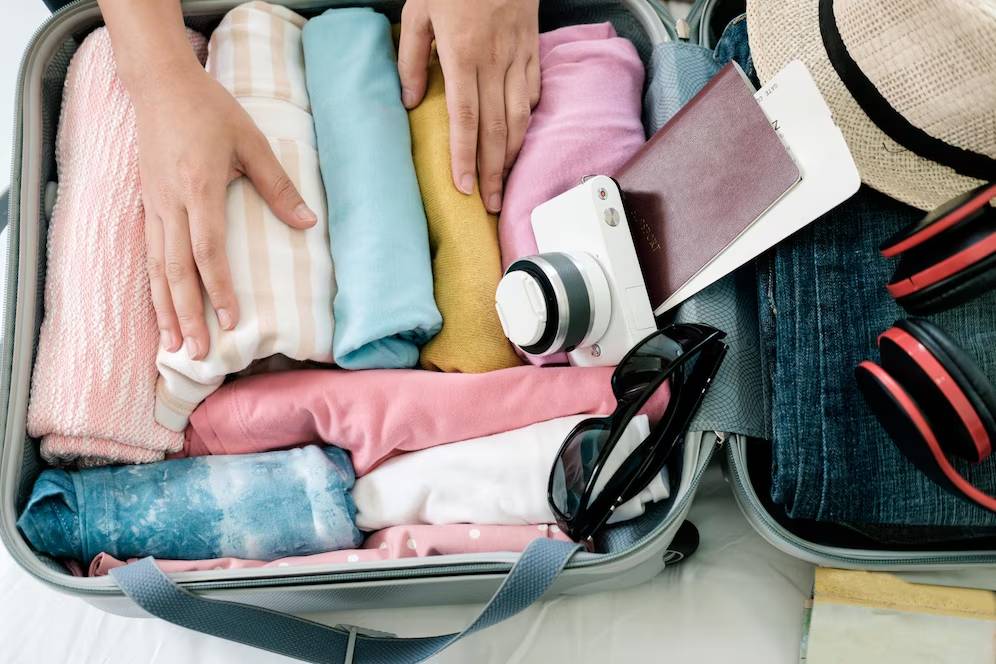 A top-down view of a packed suitcase with neatly rolled clothes, a camera, sunglasses, a passport, and a straw hat.