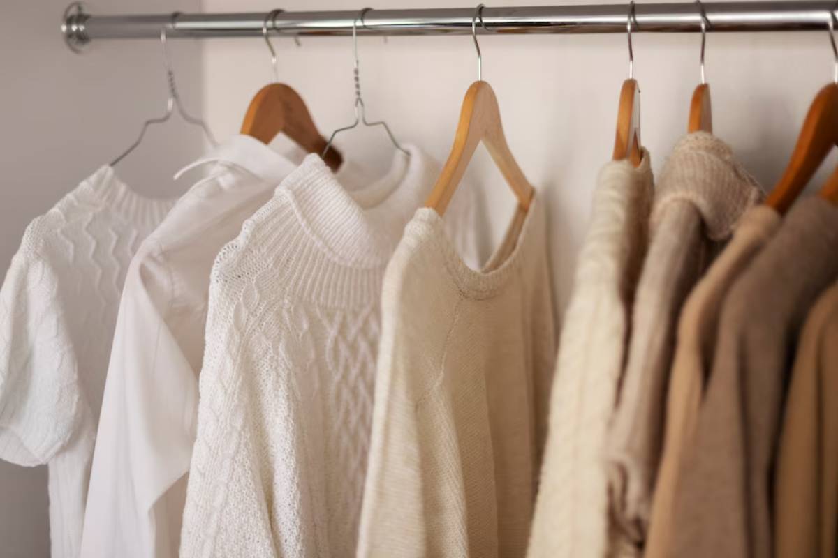 A collection of light-colored sweaters and a shirt hanging on wooden hangers, arranged neatly on a chrome rack.