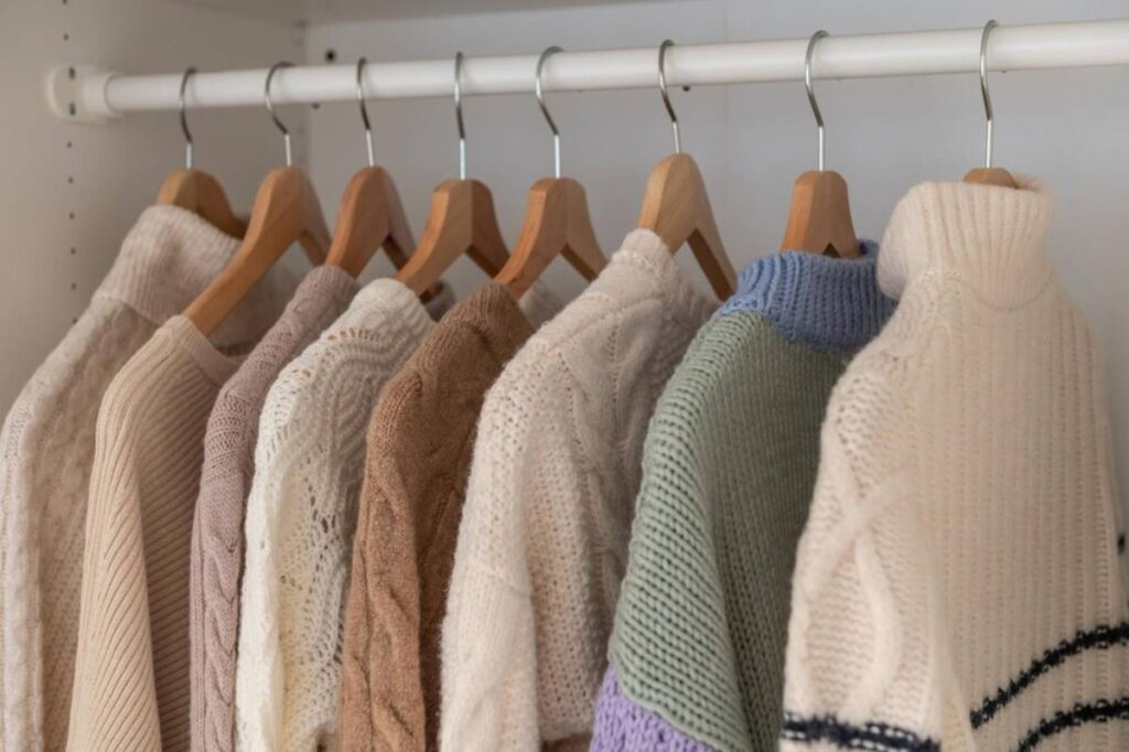 A collection of cozy, textured sweaters in soft pastel and neutral colors hanging on wooden hangers in a tidy wardrobe.