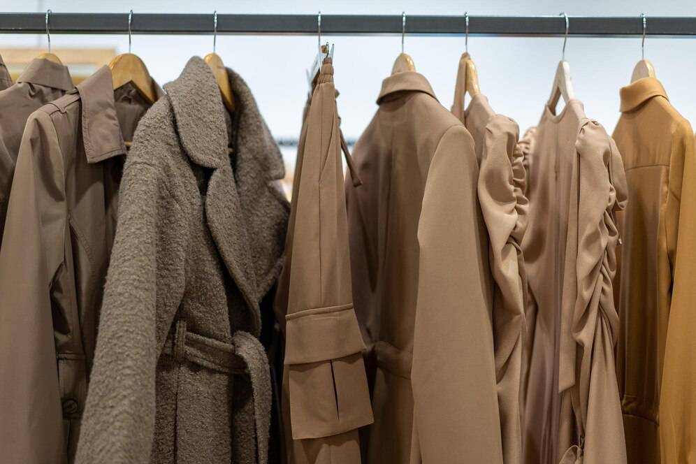 A collection of stylish beige and brown garments hangs on a rack, showcasing various textures and designs, including coats and dresses.
