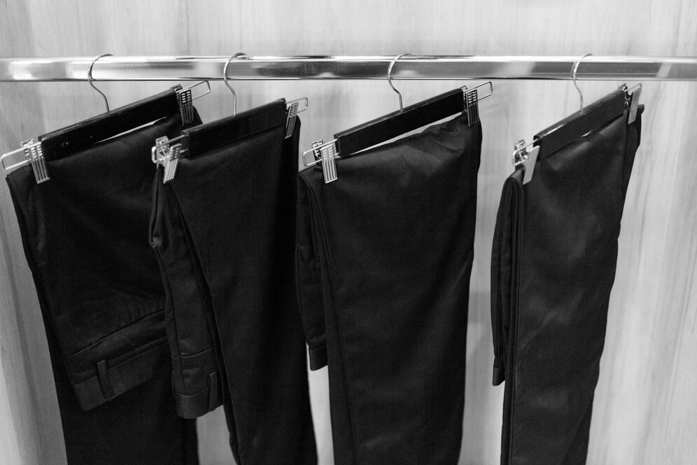 Four pairs of black trousers hanging on clips from a metal rack, against a light wooden background.