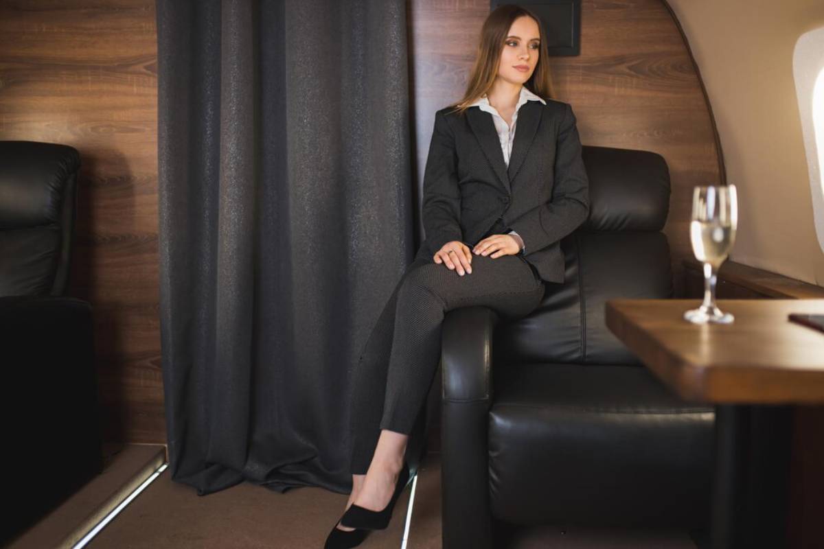 A woman in a stylish suit sits elegantly in a private jet's leather seat, with a glass of champagne on the table beside her.