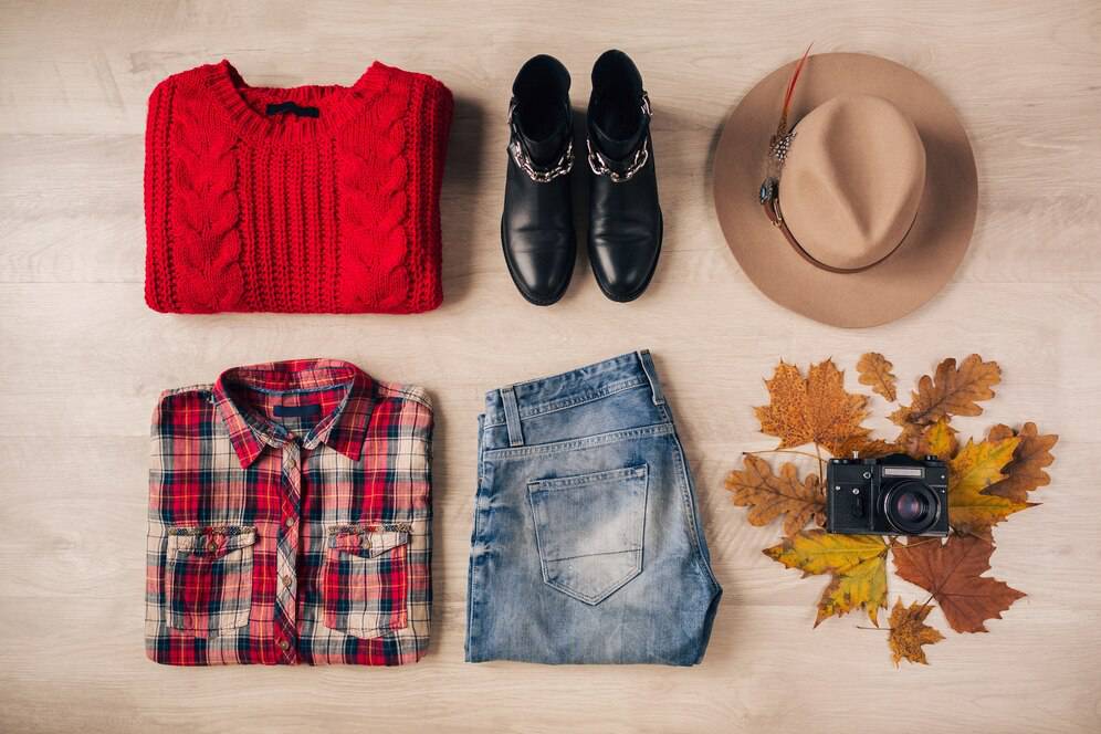 Flat lay of autumn outfit: red knit sweater, plaid shirt, jeans, black boots, tan hat, camera, and colorful leaves.