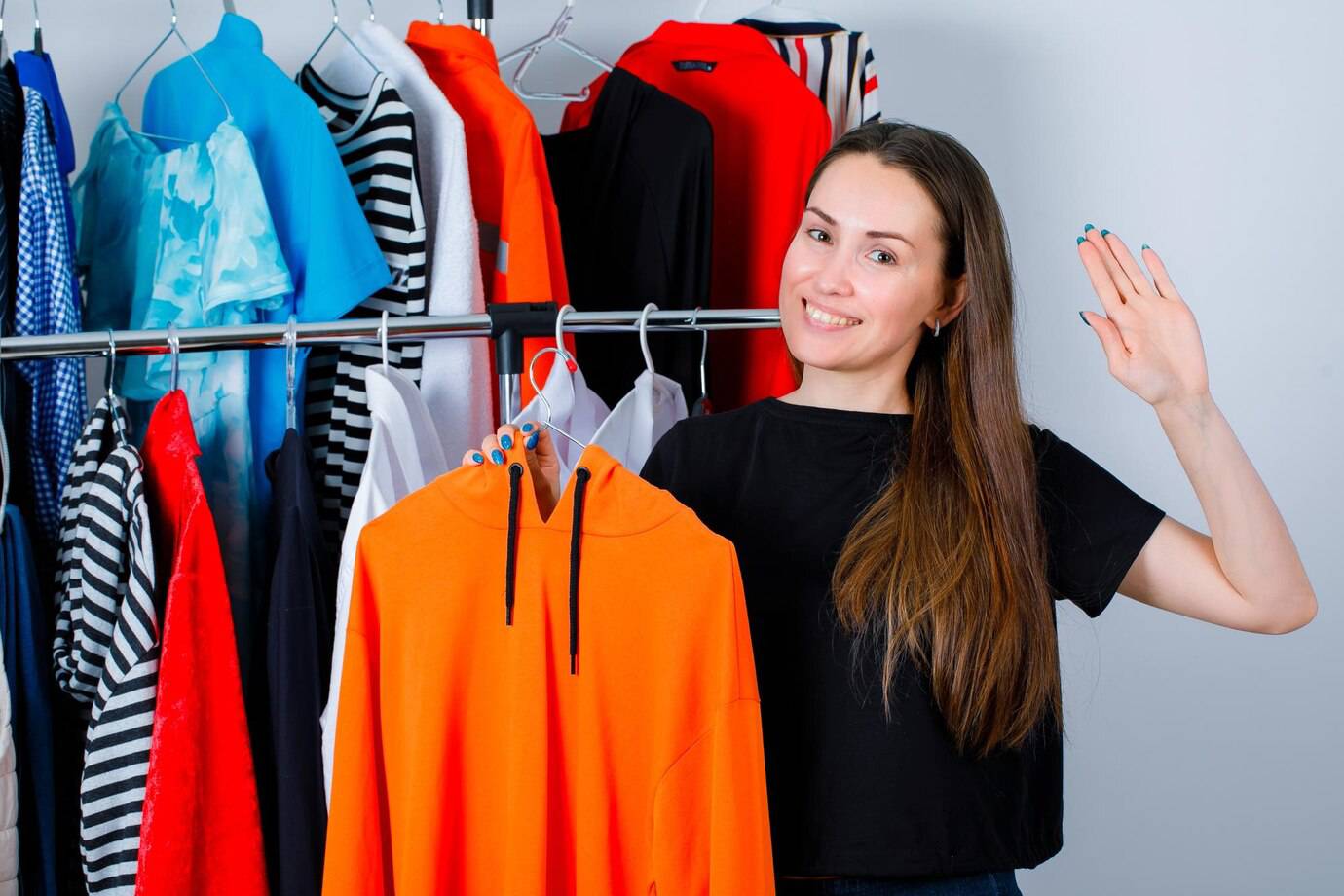 A woman in a black shirt holds up an orange hoodie in front of a clothing rack filled with colorful garments.