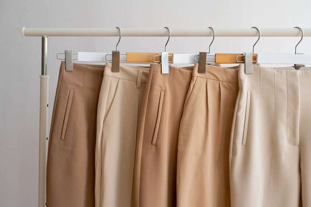 A row of stylish beige and taupe trousers on hangers, showcasing various shades of neutral fabric against a minimalist background.