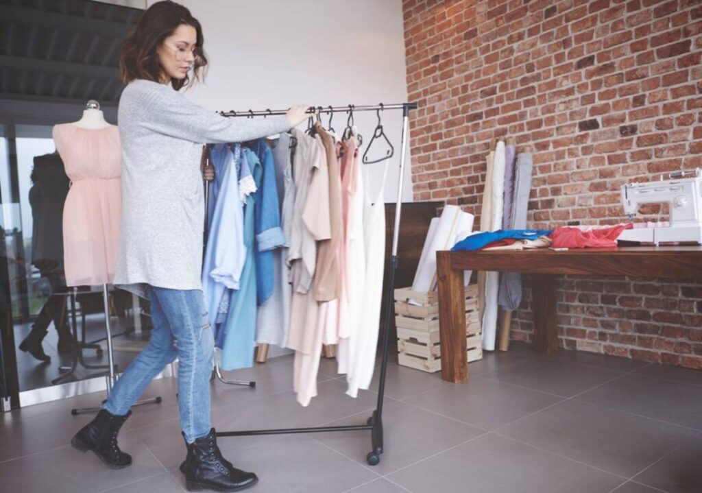 A person in a gray sweater examining clothes on a rack in a stylish studio with a brick wall and a sewing machine on the table.