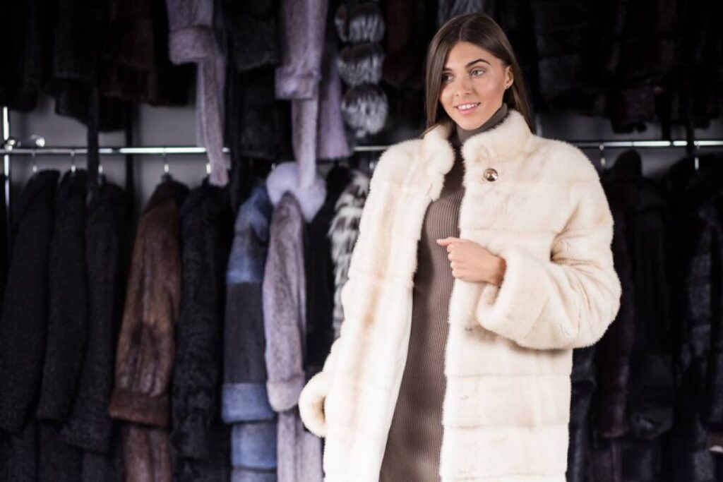 A woman models a luxurious white fur coat in front of a rack filled with various colored fur coats, showcasing a stylish collection.