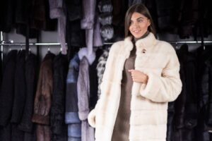 A woman models a luxurious white fur coat in front of a rack filled with various colored fur coats, showcasing a stylish collection.