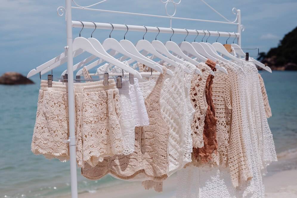 A rack displaying light, beach-style clothing in neutral tones hanging by the sea, with waves and rocks in the background.