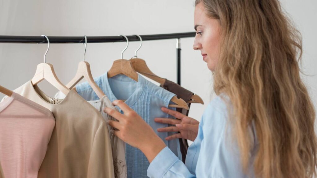 A person with long hair examines a rack of clothing, which includes various tops in soft colors and fabrics.