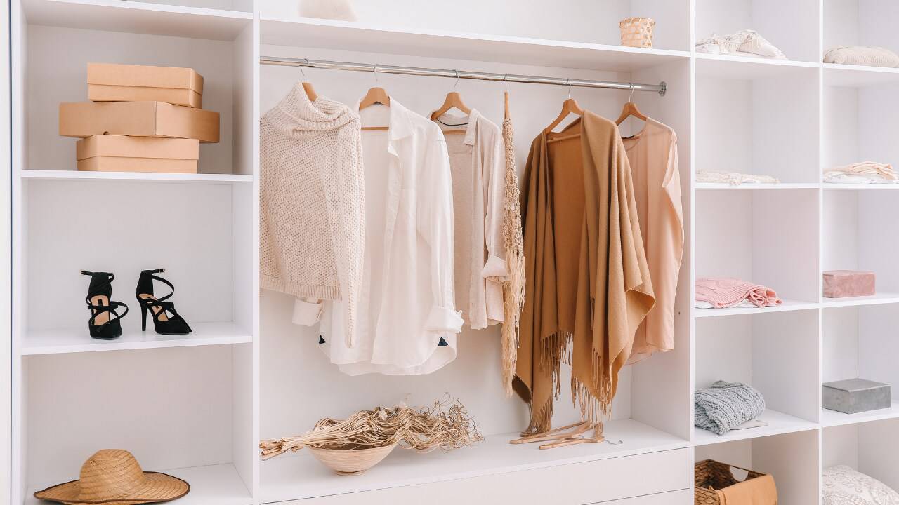 A neatly organized closet featuring beige and tan clothing on hangers, black high heels, decorative boxes, and a straw hat.