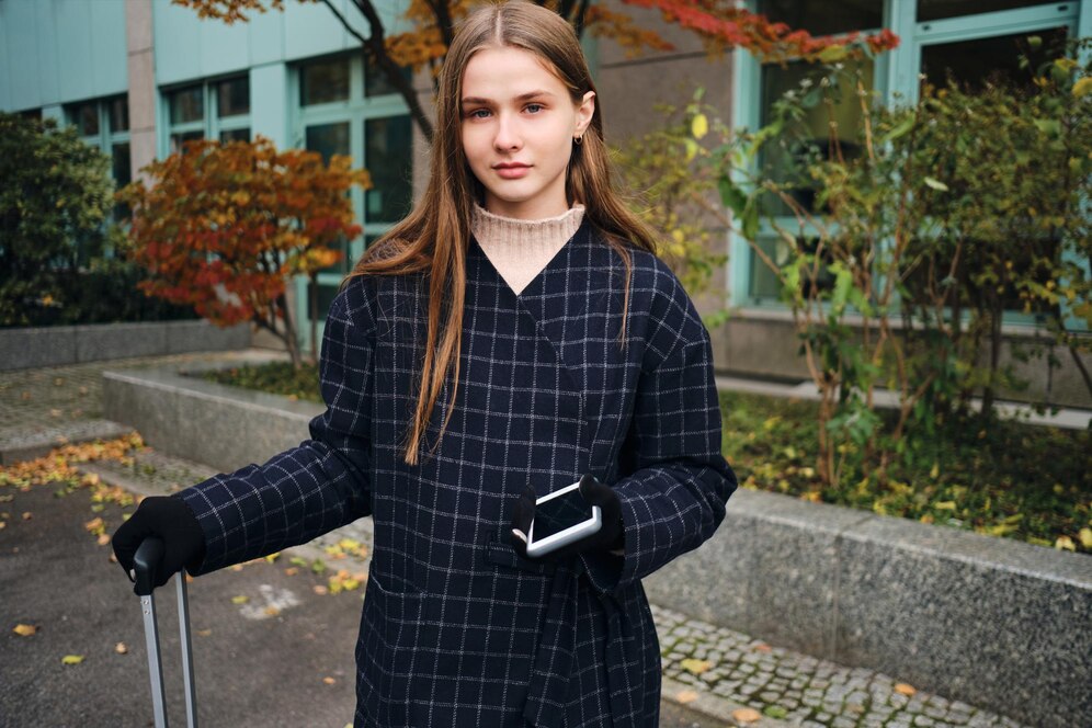 A person wearing a dark checkered coat stands outdoors, holding a smartphone and pulling a suitcase, surrounded by autumn foliage.