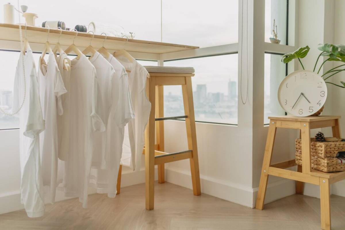 A bright room featuring white shirts hanging by a window, a wooden stool, and a clock atop a small table with a woven basket.