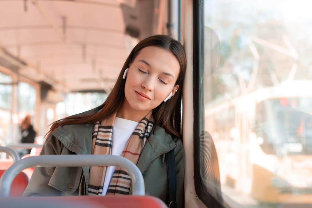 A young woman rests her head against a bus window, wearing a jacket and a patterned scarf, while a city street is visible outside.