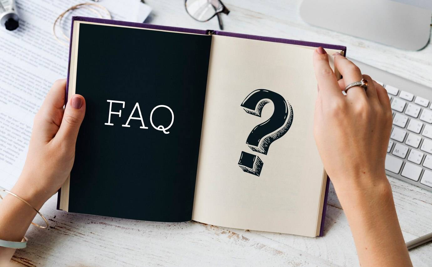 A person holding an open book with  on one page and a large question mark on the opposite page, with a workspace in the background.