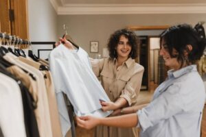 Two women examine a light blue shirt while standing in a clothing store, surrounded by various garments on display.