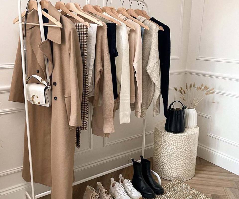 A stylish clothing rack displays various neutral-toned outfits, with boots and sneakers on the floor, beside decorative vases.