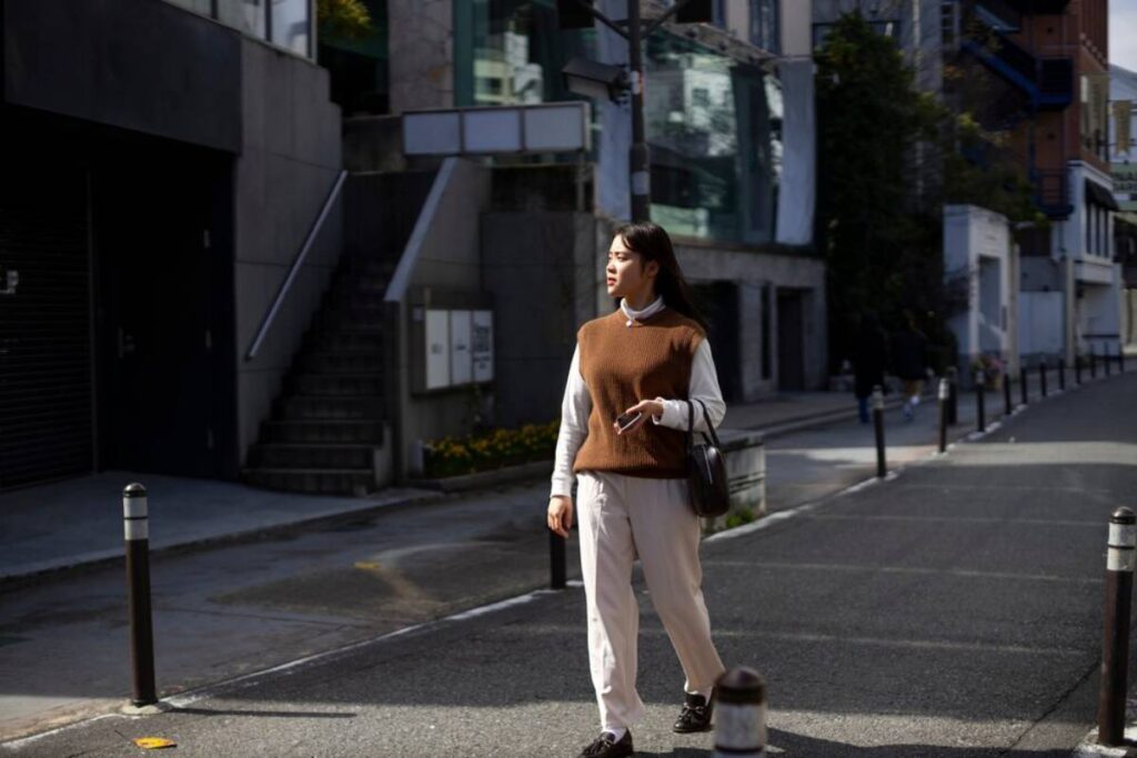 A woman walks down a city street, wearing a brown vest over a white shirt and beige trousers, holding a phone and a handbag.