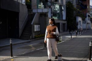 A woman walks down a city street, wearing a brown vest over a white shirt and beige trousers, holding a phone and a handbag.