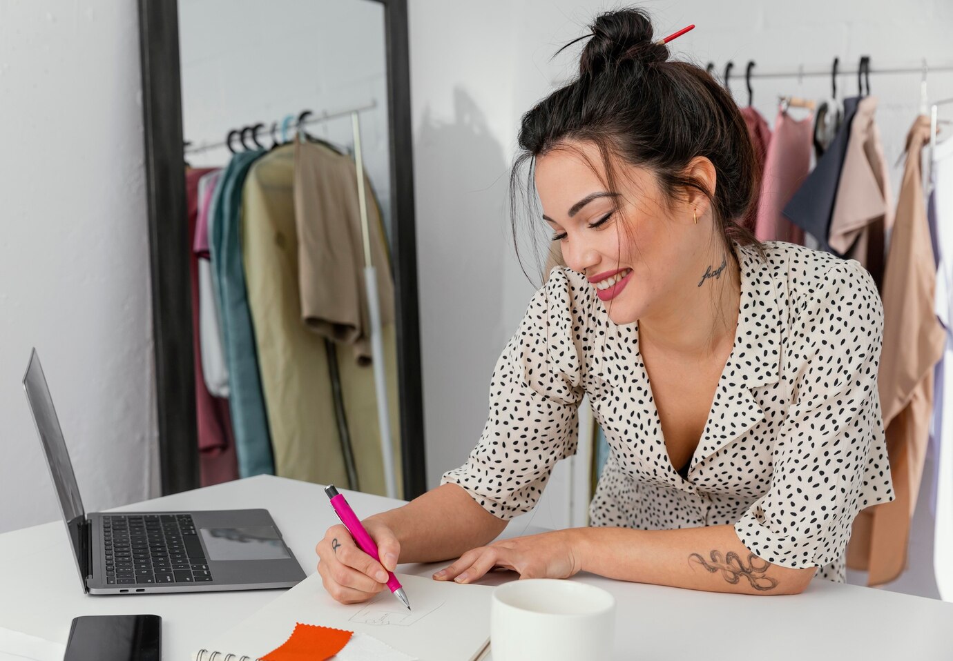 A woman wearing a polka-dot blouse sketches in a notebook with a pink pen