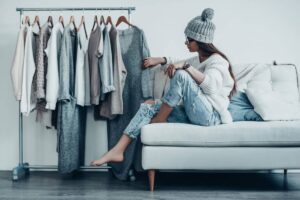 A person in a cozy sweater and beanie sits on a white couch, surrounded by a rack of stylish, neutral-toned clothing.
