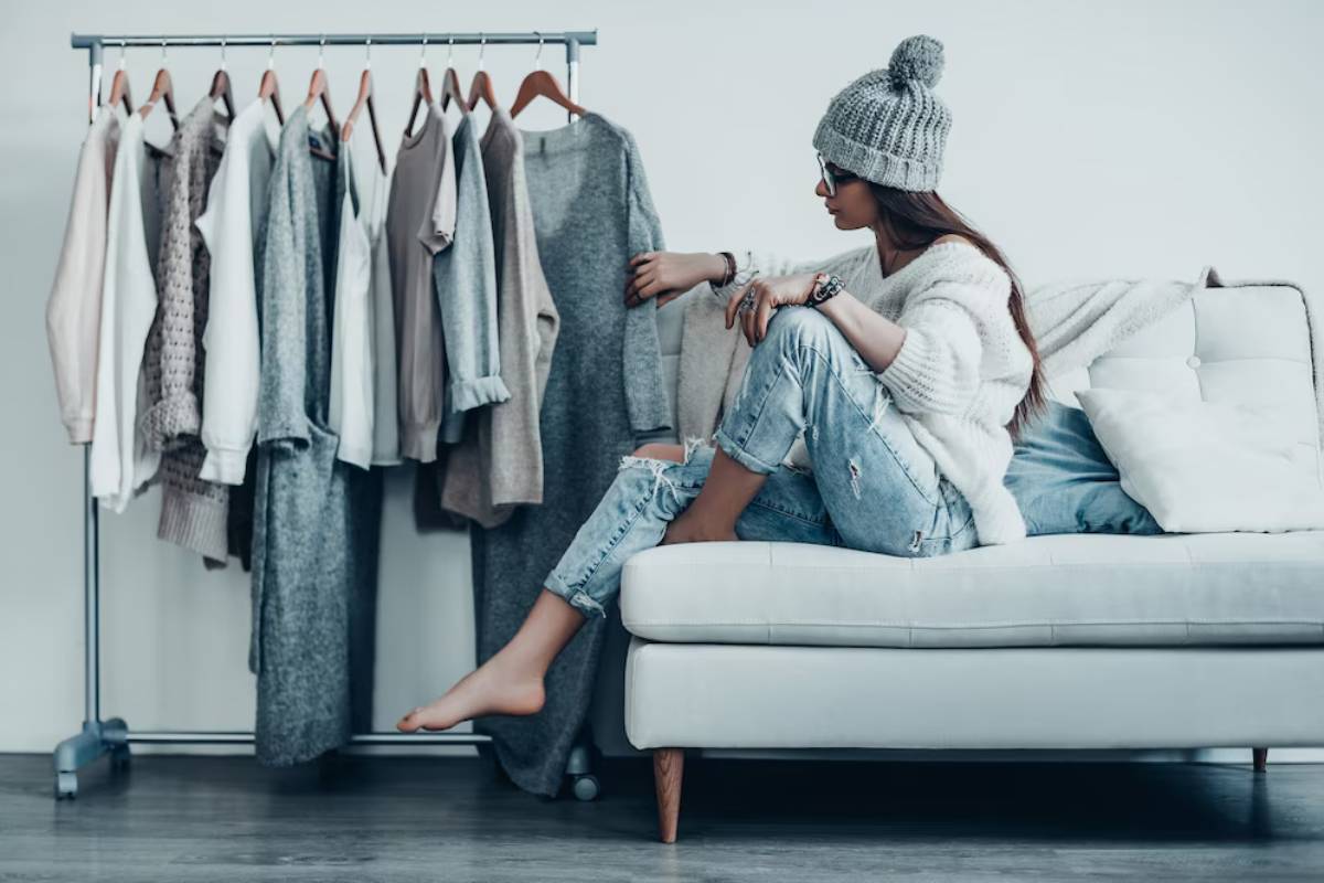 A person in a cozy sweater and beanie sits on a white couch, surrounded by a rack of stylish, neutral-toned clothing.