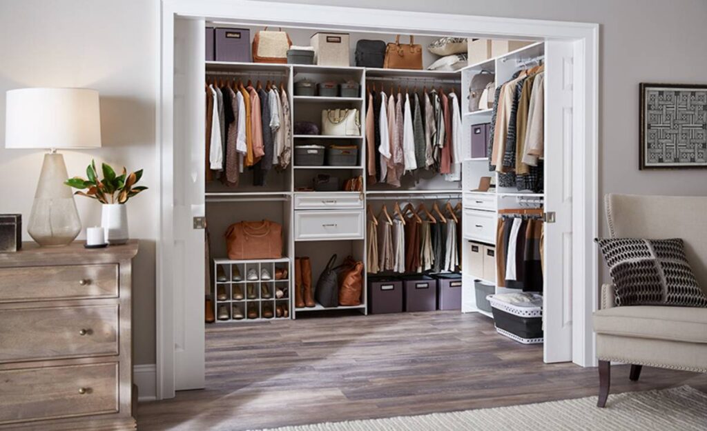 A stylish organized closet with hanging clothes, folded items, and handbags alongside a dresser and a cozy chair in a modern room.
