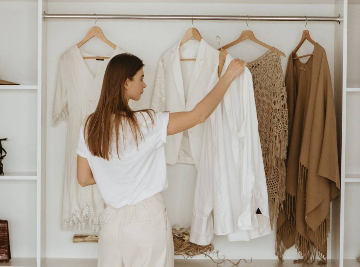 A woman stands in front of a well-organized wardrobe, selecting a white shirt from a variety of hanging garments.