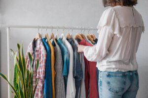 A woman in a white blouse browses a rack of colorful shirts, next to a green plant, against a light gray background.