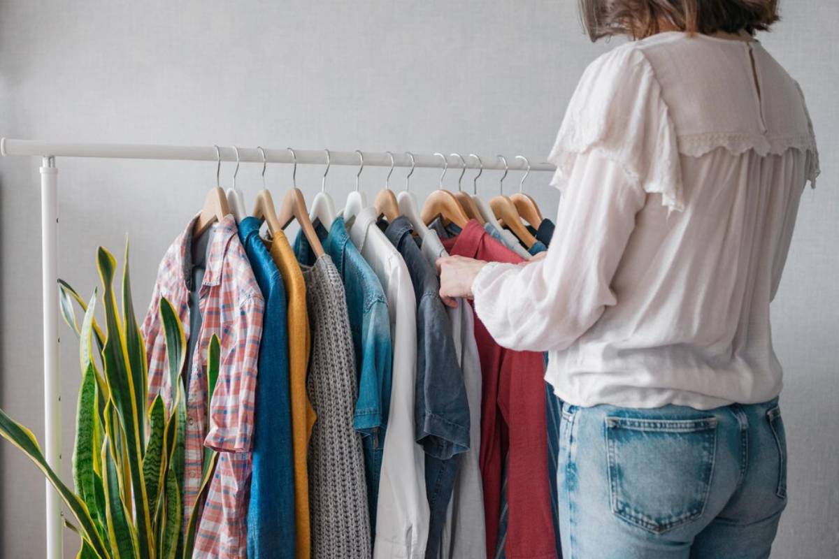 A woman in a white blouse browses a rack of colorful shirts, next to a green plant, against a light gray background.