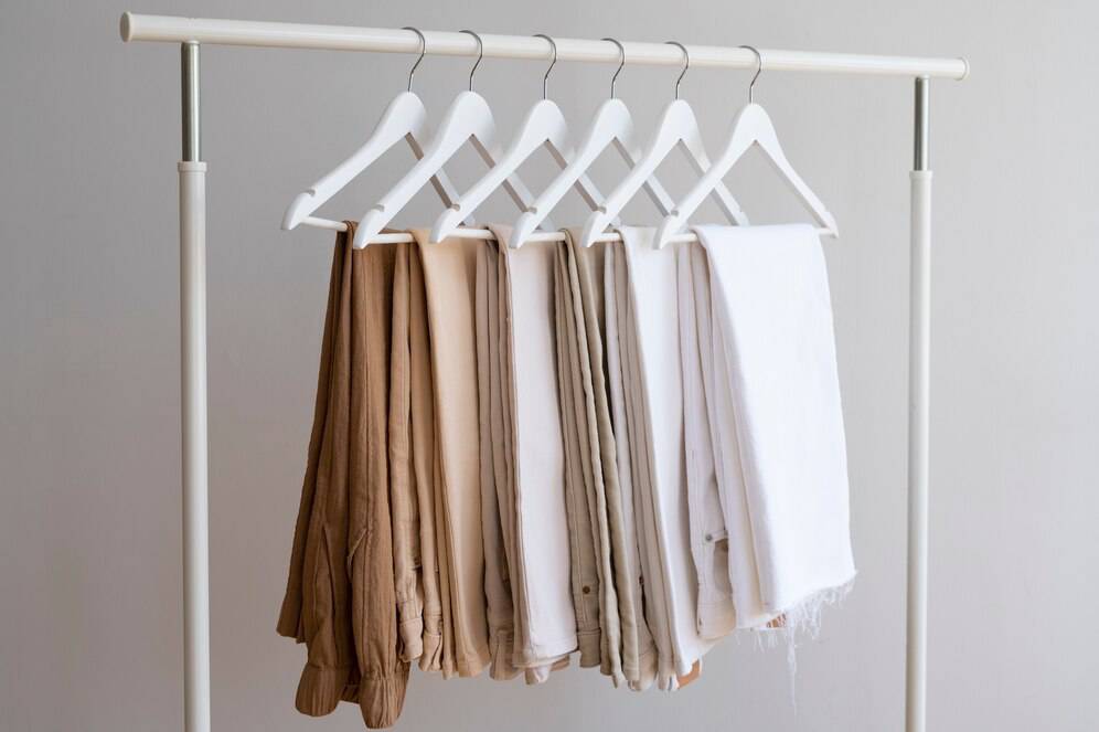 A minimalist clothing rack displays a gradient of neutral-toned pants and fabrics, neatly hung on white hangers against a light backdrop.