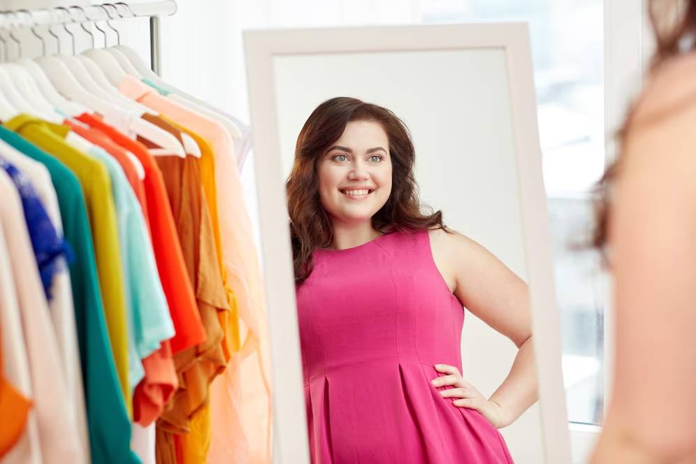A person in a pink dress poses confidently in front of a mirror, surrounded by colorful clothing on hangers.