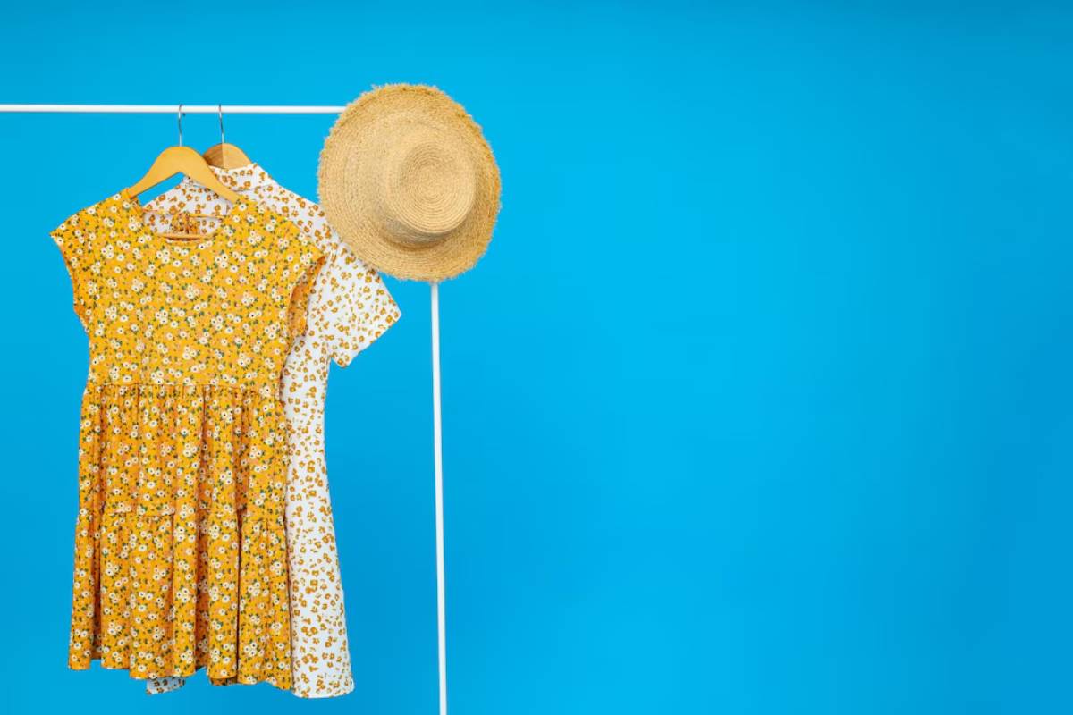 A vibrant yellow floral dress, a white floral dress, and a straw hat hanging on a display against a bright blue background.