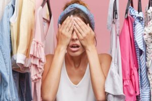 A person sits surrounded by colorful clothing on hangers, appearing overwhelmed while holding their head in their hands.