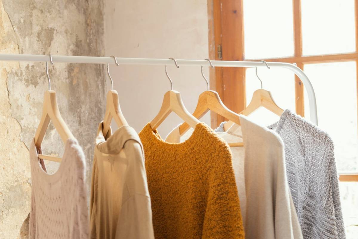 Five cozy sweaters hang on a white rack, illuminated by natural light from a window, against a textured wall.