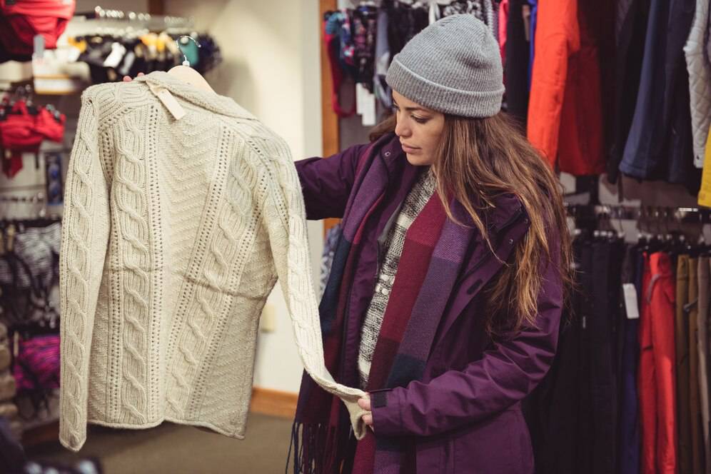 A woman in a purple jacket and gray beanie holds up a thick, cream-colored knitted sweater in a retail store.